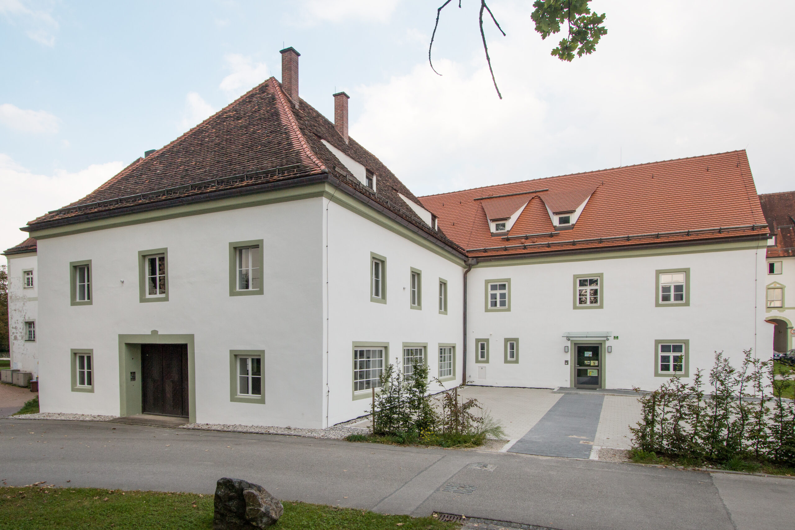 Ein großes, weißes, zweistöckiges Gebäude, das an die Hochschule Coburg erinnert, verfügt über ein rotes Ziegeldach und zahlreiche Fenster. Es liegt auf einer Rasenfläche mit gepflastertem Eingang und ist von vereinzelten Bäumen und Sträuchern umgeben.