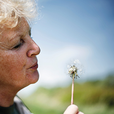 Eine Person mit lockigem, hellem Haar steht im Außenbereich der Hochschule Coburg und bläst sanft in eine Pusteblume. Der blaue Himmel und der leicht unscharfe Hintergrund rufen die Essenz eines sonnigen Tages hervor und fangen einen Moment heiterer Schönheit auf dem Campus ein.