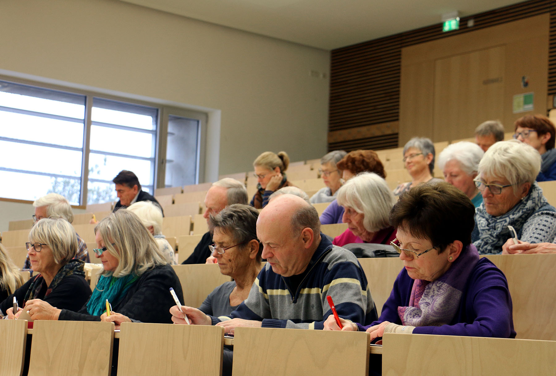 In einem Hörsaal der Hochschule Coburg sitzen ältere Erwachsene an ihren Schreibtischen und machen sich aufmerksam Notizen. Der Raum verfügt über große Fenster und helle Holzmöbel, was eine fleißige und engagierte Atmosphäre schafft.