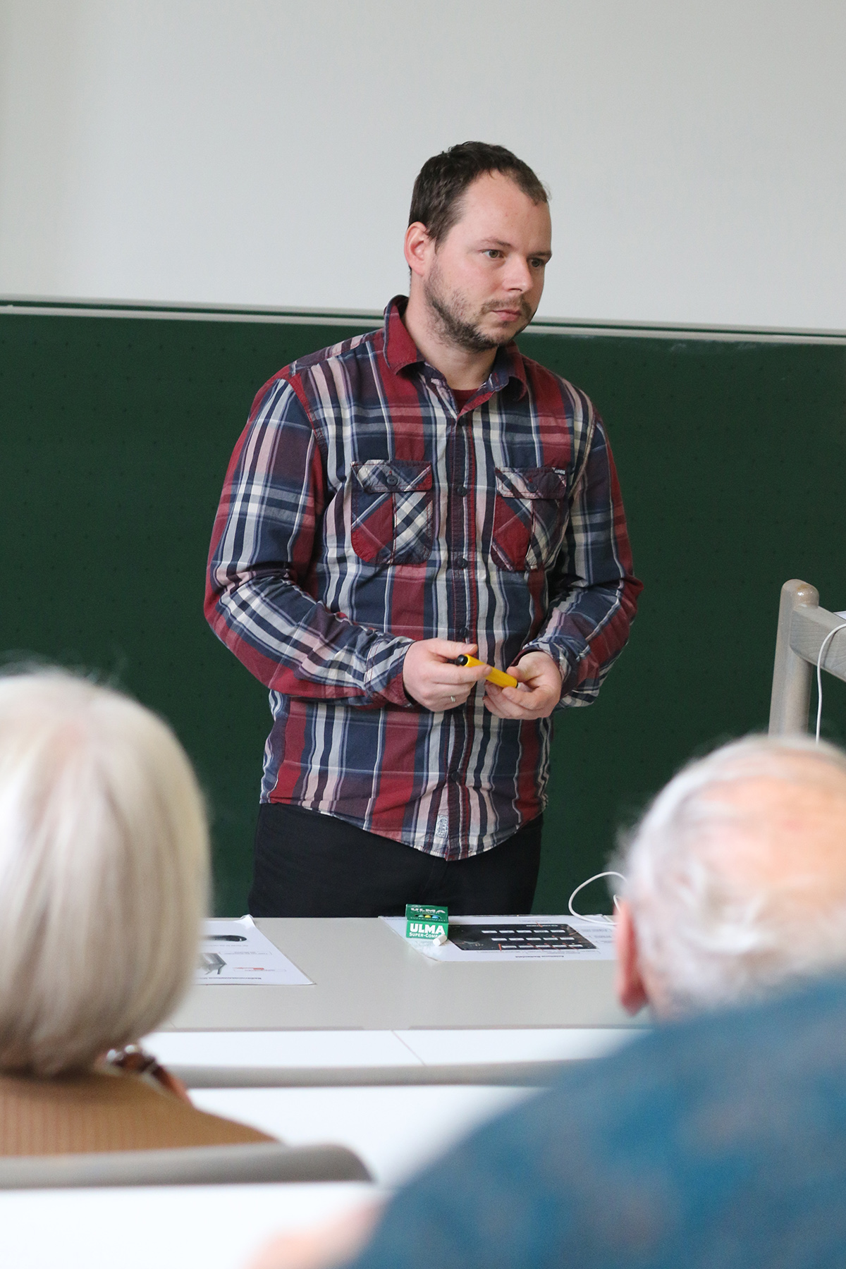 Ein Mann im karierten Hemd steht vor einem Hörsaal der Hochschule Coburg. Er hält einen Stift und eine Packung Kaugummi in der Hand. Er scheint eine Vorlesung oder einen Vortrag zu halten. Im Vordergrund sind verschwommene Köpfe aufmerksamer Zuhörer zu erkennen.