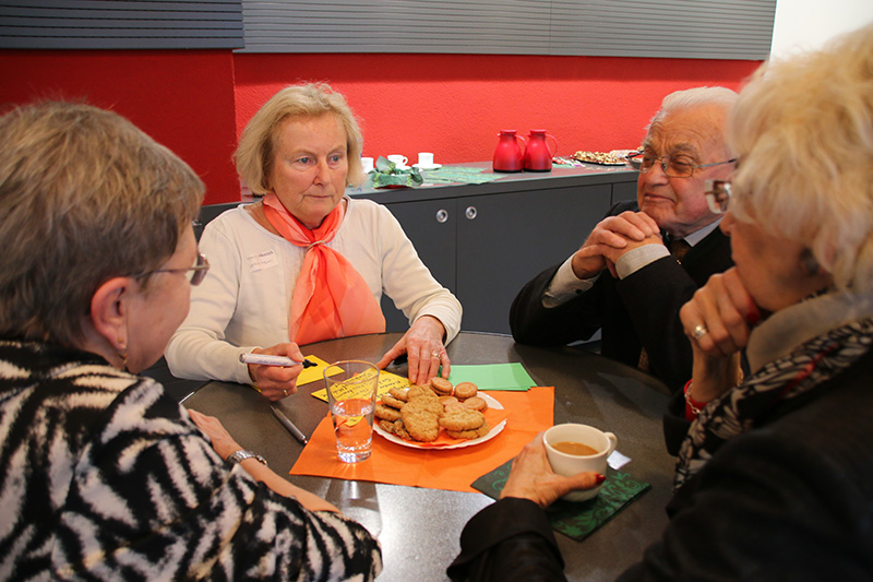 Eine Gruppe von vier älteren Menschen sitzt mit einem Teller Kekse um einen Tisch und genießt Kaffee oder Tee. Der Raum mit seinen roten und grauen Wänden wirkt warm. Eine Person hält einen Stift in der Hand, während alle sich angeregt unterhalten und vielleicht an ihre Tage an der Hochschule Coburg zurückdenken.