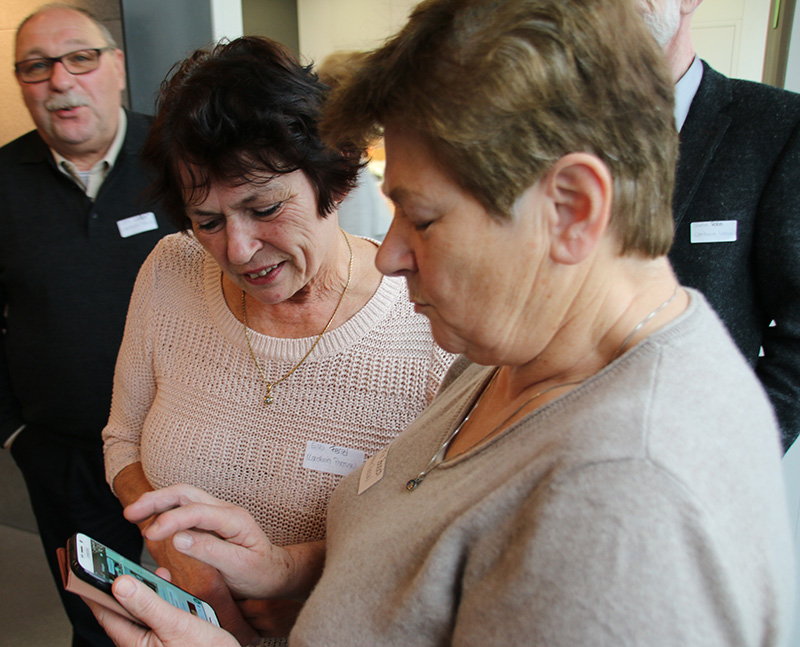 Zwei Frauen stehen dicht beieinander und schauen auf ein Smartphone, das eine in der Hand hält, während sie sich unterhalten. Im Hintergrund sind zwei Männer zu sehen, die teilweise zu sehen sind. Alle tragen Namensschilder bei einer Veranstaltung der Hochschule Coburg und scheinen sich in einem geselligen Umfeld in Innenräumen aufzuhalten.