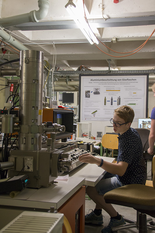 Eine Person mit Brille bedient ein großes wissenschaftliches Instrument in einem Labor der Hochschule Coburg. Sie sitzt da und arbeitet konzentriert an den Bedienelementen. Hinter ihr hängt ein Poster mit Texten und Diagrammen, während die Umgebung mit verschiedenen Geräten und Kabeln vollgestopft ist.