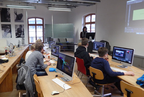 In einem Hörsaal der Hochschule Coburg sitzen die Studierenden an Tischen mit Apple-Computern. Ein Lehrer steht neben Whiteboard und Projektionsleinwand und leitet die Sitzung. Der Raum wird durch große Fenster erhellt und mit Schwarzweißfotos geschmückt.