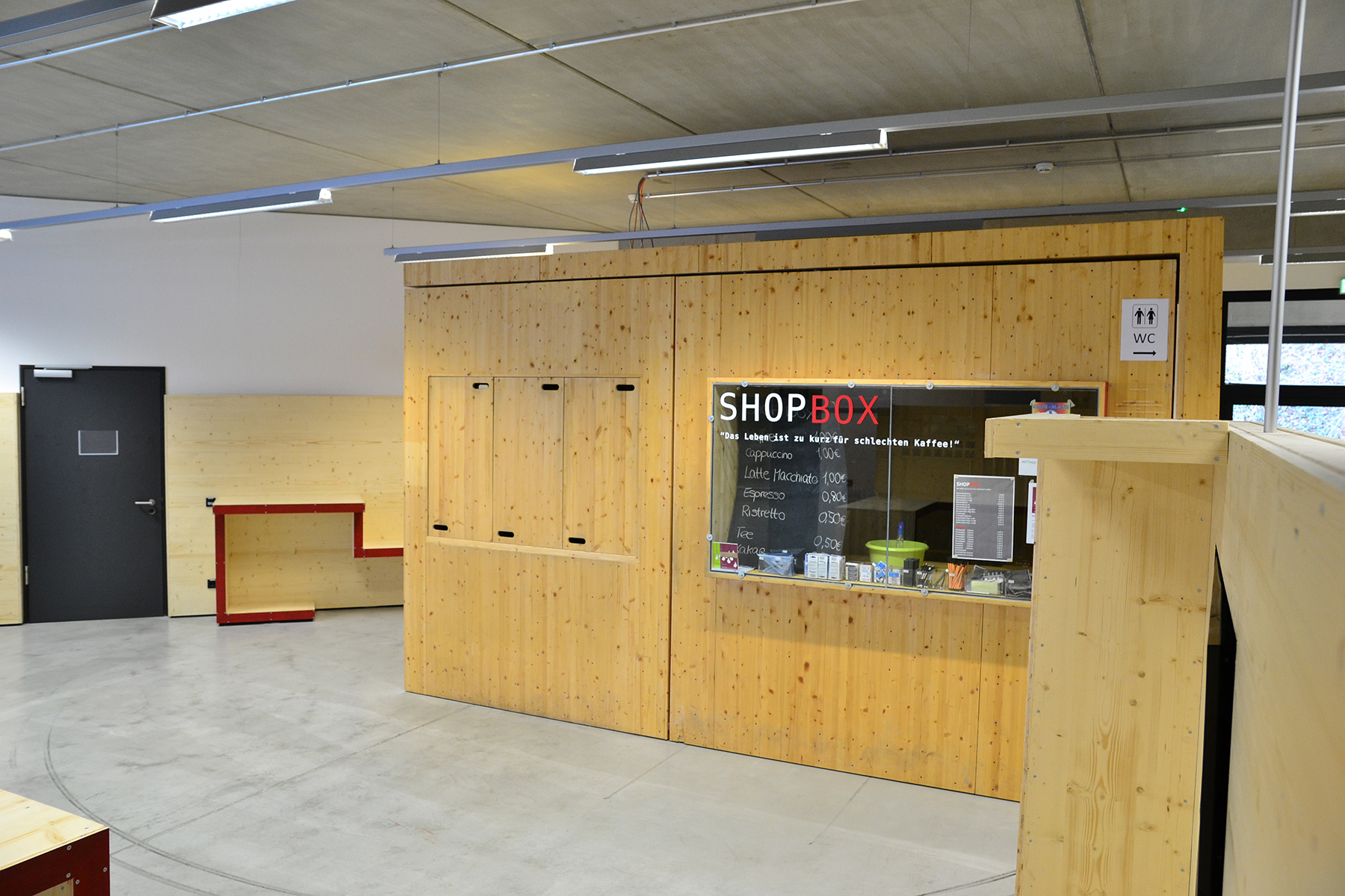 Ein moderner Innenraum der Hochschule Coburg besticht durch helle Holzvertäfelungen und einen „SHOP BOX“-Schalter mit Preisauskünften. Ein kleines schwarzes Schild weist auf die Toiletten hin und betont den Betonboden und das minimalistische Design des Raums.