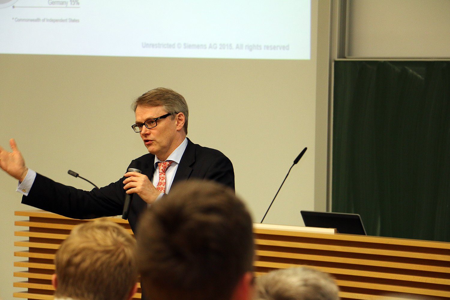 Ein Mann im Anzug spricht in ein Mikrofon und gestikuliert mit der anderen Hand in einem Hörsaal der Hochschule Coburg. Im Hintergrund sind eine Projektionsleinwand und ein Rednerpult zu sehen, im Vordergrund sind aufmerksame Zuhörer zu sehen.