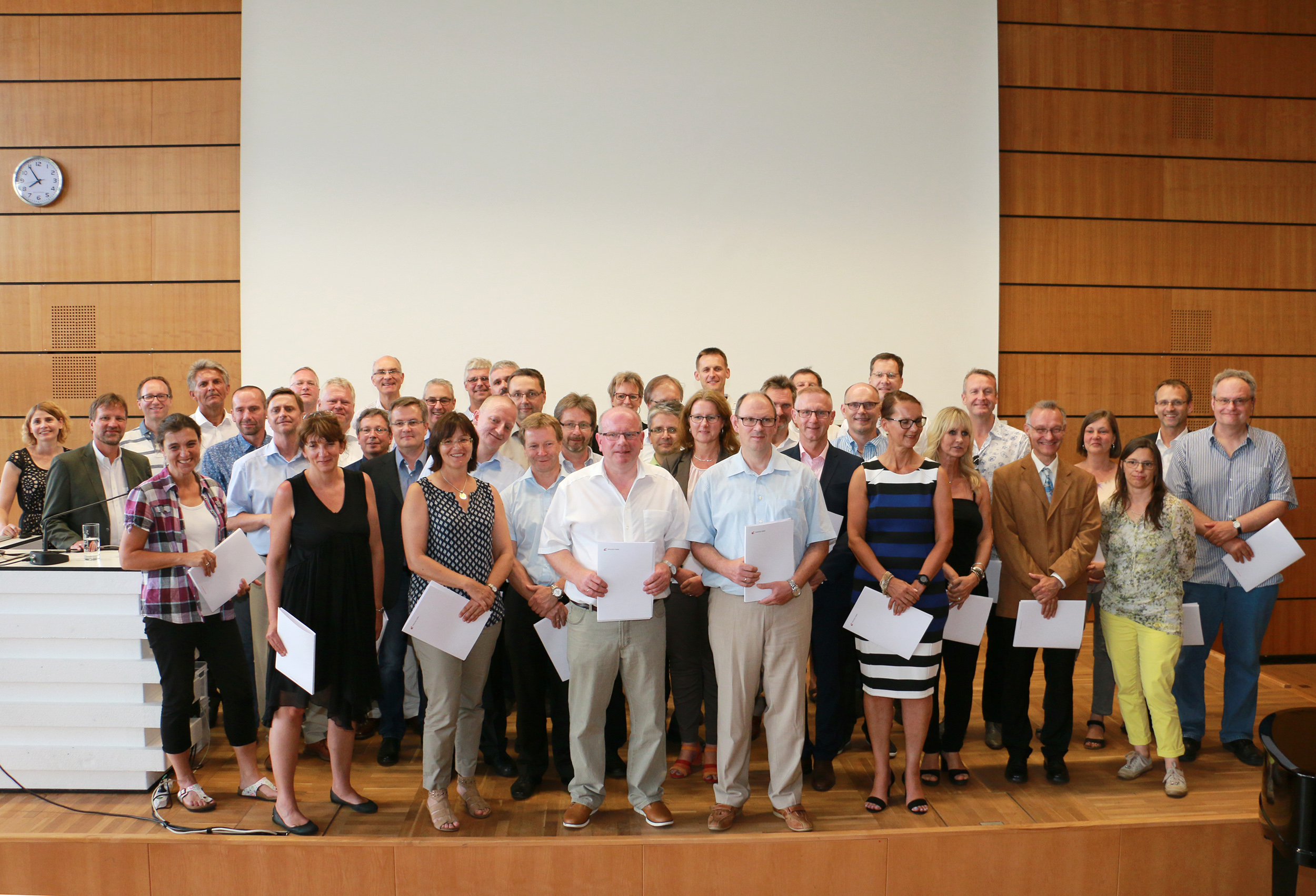 Eine Gruppe von Mitarbeitern der Hochschule Coburg posiert für ein Foto in einem holzgetäfelten Raum mit einer weißen Leinwand im Hintergrund. Einige halten Papiere in den Händen. Die Gruppe besteht aus Männern und Frauen, alle in legerer Business-Kleidung gekleidet. Eine Uhr an der Wand zeigt 2:10 Uhr.