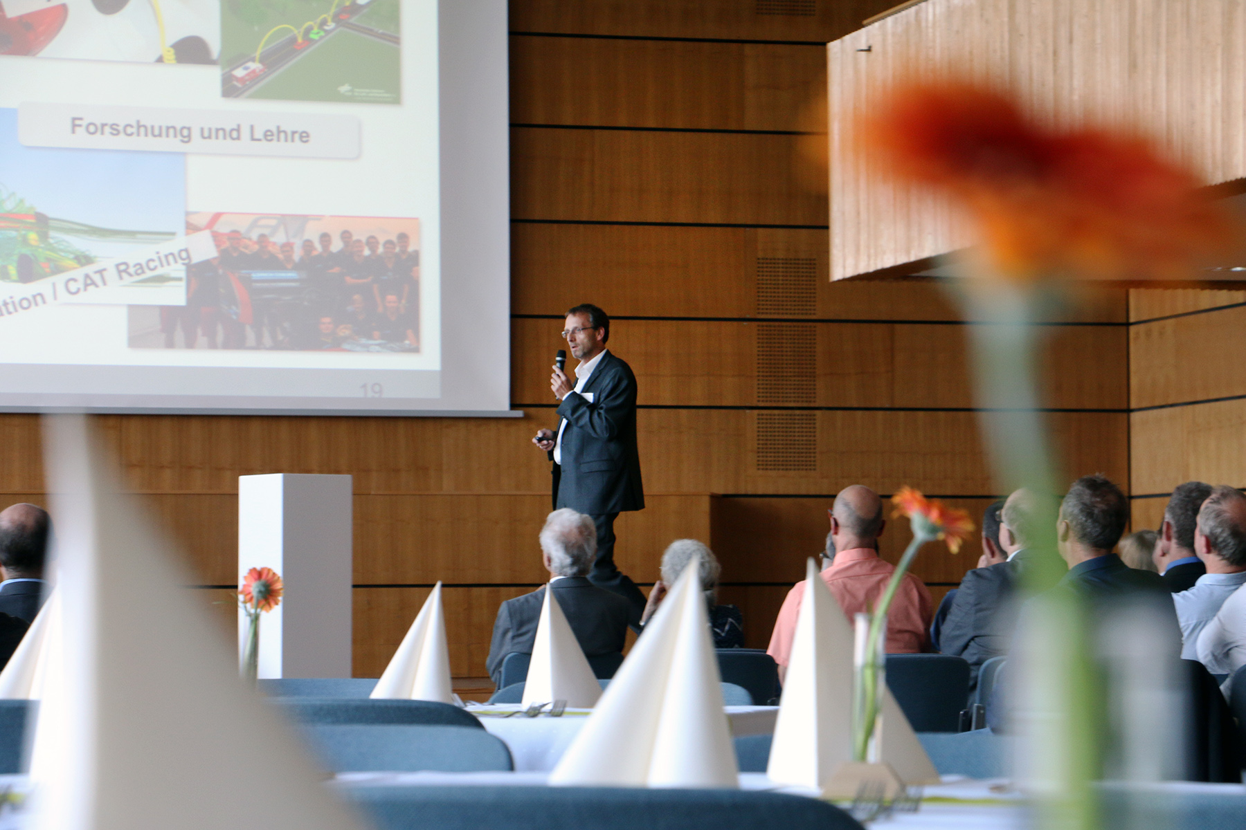 Eine Person im Anzug steht auf einer Bühne bei der Konferenz der Hochschule Coburg und hält einen Vortrag. Die Zuschauer sitzen an Tischen, die mit weißen Tischdecken und spitzen Servietten geschmückt sind. Eine orangefarbene Blume ziert den Vordergrund, ist teilweise unscharf und verleiht der Kulisse einen Hauch von Eleganz.