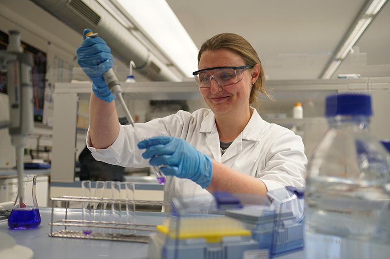 Eine Wissenschaftlerin der Hochschule Coburg, mit Schutzbrille und Laborkittel, benutzt im Labor eine Pipette. Sie trägt blaue Handschuhe und ist von einer Reihe von Laborgeräten und Flaschen umgeben.