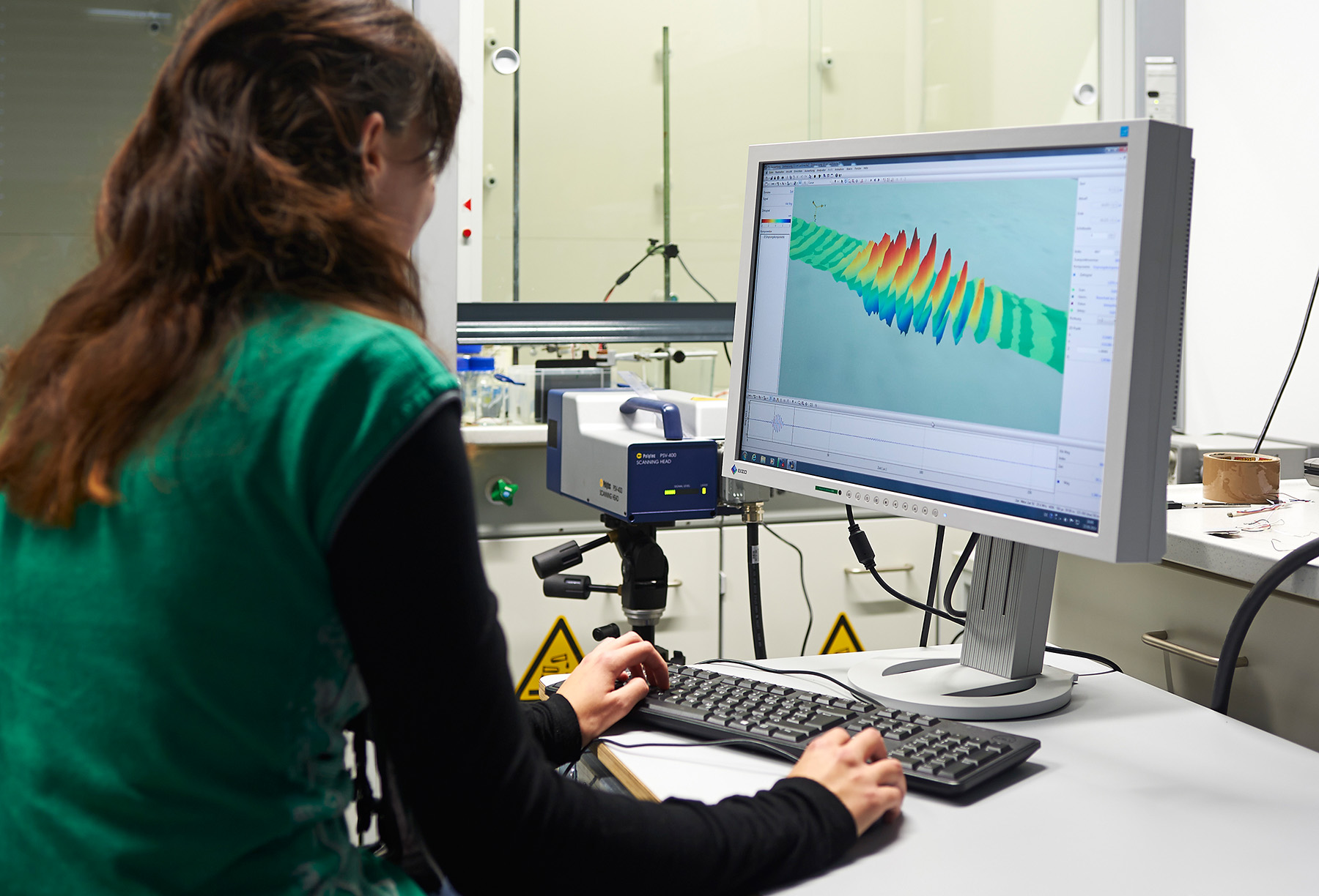 Eine Person im grünen Hemd sitzt an einem Schreibtisch und bedient einen Computer im Labor der Hochschule Coburg. Der Monitor zeigt farbenfrohe 3D-Grafiken oder Datenvisualisierungen, während daneben ein wissenschaftliches Instrument steht. Im Hintergrund sind verschiedene Laborgeräte zu sehen.