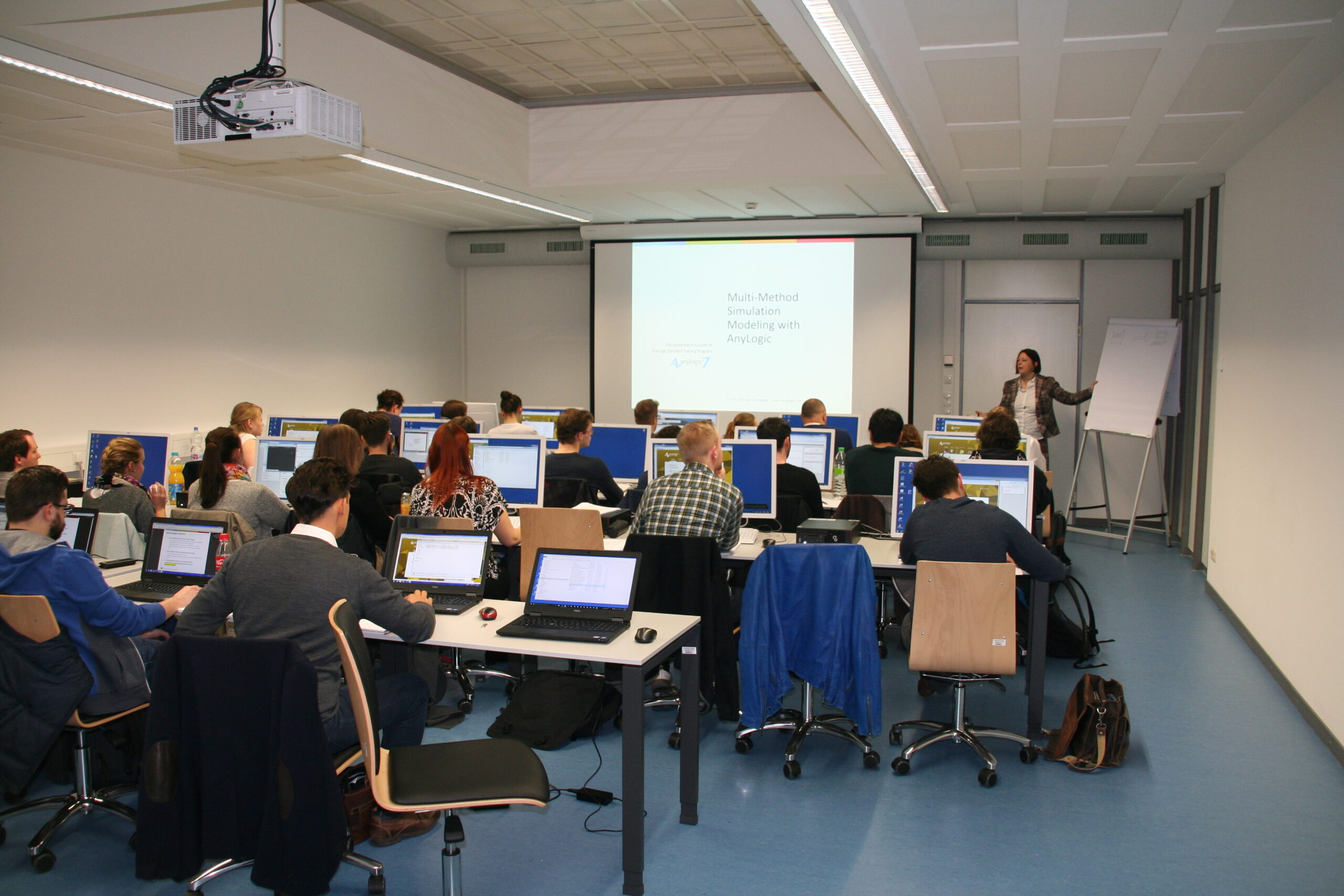 In einem modernen, professionellen Umfeld der Hochschule Coburg sitzen Studierende mit Laptops an Schreibtischen. Ein Vortragender steht vorne neben einem Bildschirm, auf dem eine Präsentation zum Thema „Multimethodische Simulationsmodellierung mit AnyLogic“ läuft.