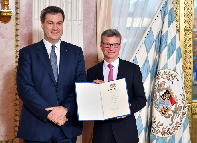 Zwei Männer in Anzügen stehen in einem formellen Raum der Hochschule Coburg. Einer der Männer hält lächelnd ein Zertifikat in der Hand. Neben ihnen ist eine dekorative Flagge mit einem heraldischen Emblem zu sehen, was darauf hinweist, dass sie sich auf einer offiziellen Veranstaltung oder Zeremonie befinden.