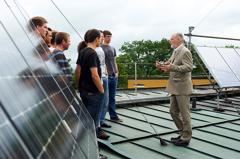 Eine Gruppe junger Erwachsener der Hochschule Coburg steht auf einem Dach mit Solarmodulen und hört einem älteren Mann im Anzug aufmerksam zu, der beim Sprechen gestikuliert. Die Außenszene wird durch im Hintergrund sichtbare Bäume bereichert.