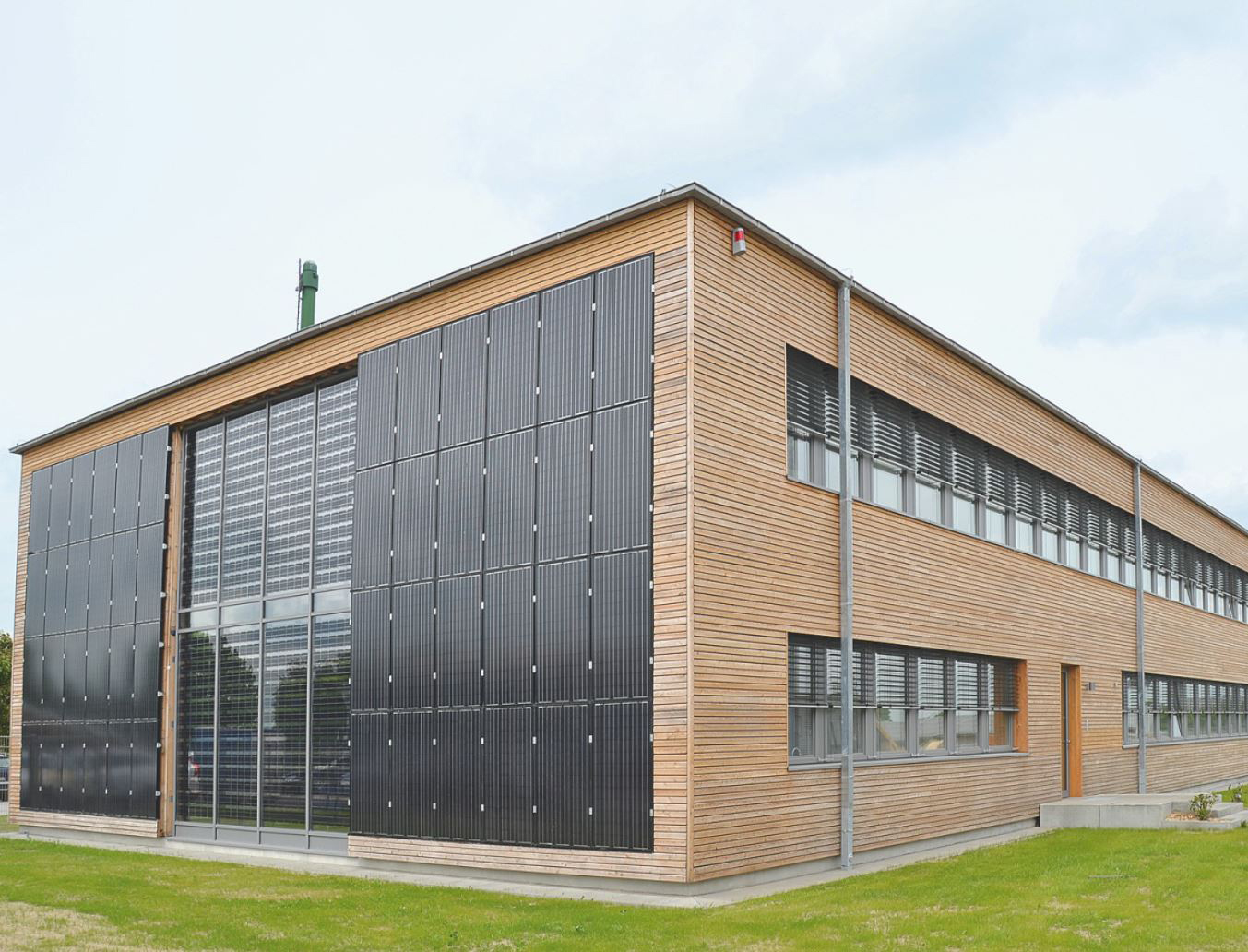 Ein modernes zweistöckiges Gebäude mit Holzverkleidung und großen Solarmodulen, das an die nachhaltigen Designs der Hochschule Coburg erinnert, verfügt über große Fenster. Es liegt anmutig auf einer Rasenfläche unter einem teilweise bewölkten Himmel.