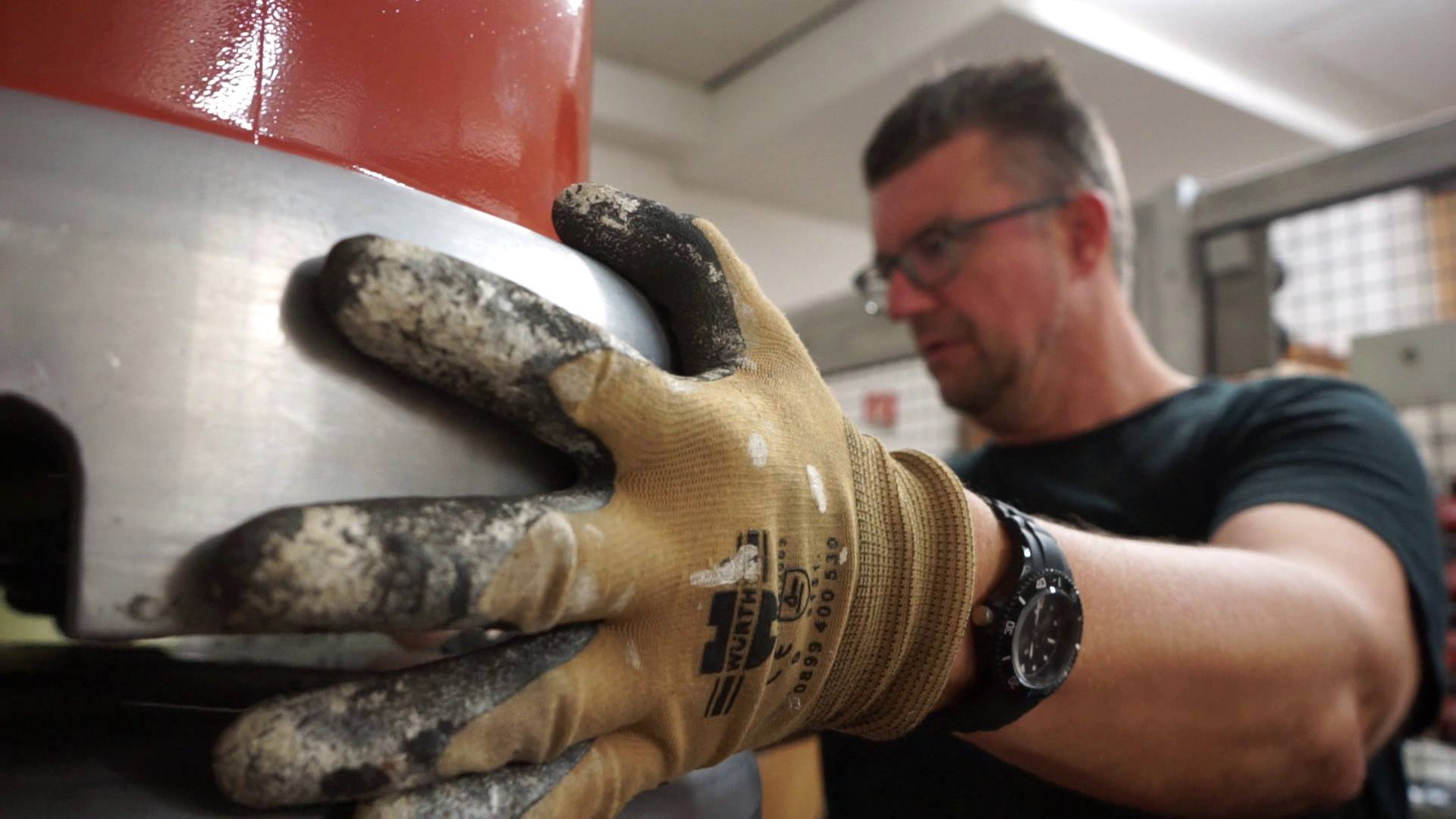 Eine Person mit Schutzhandschuhen bedient an der Hochschule Coburg konzentriert Maschinen. Die behandschuhten Hände greifen ein großes Metallobjekt, dessen rote und silberne Teile unter den Werkstattlichtern glitzern. Im Hintergrund heben moderne Industrieanlagen die hochmoderne Lernumgebung der Hochschule hervor.