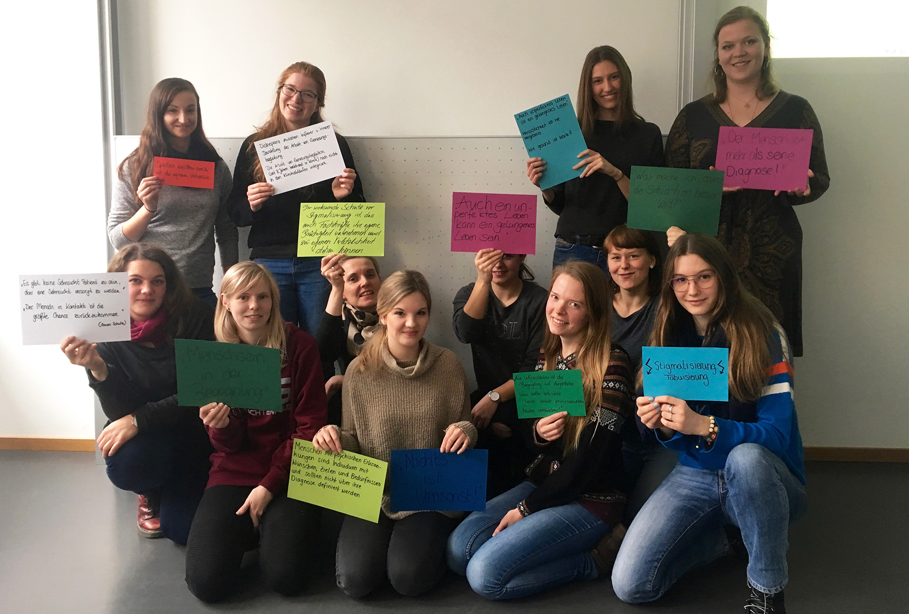 Eine Gruppe von dreizehn Personen der Hochschule Coburg lächelt und hält bunte Schilder mit Botschaften in deutscher Sprache. Sie posieren drinnen vor einer weißen Wand und schaffen so eine lockere und freundliche Atmosphäre.