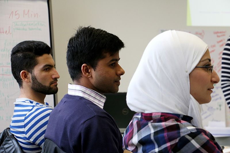 Eine Seitenansicht von drei Personen, die in einem Hörsaal der Hochschule Coburg sitzen und aufmerksam zuhören. Die Personen tragen Freizeitkleidung und eine trägt ein Kopftuch. Im Hintergrund ist eine Tafel mit Text zu sehen, die das Wesen konzentrierten akademischen Engagements einfängt.