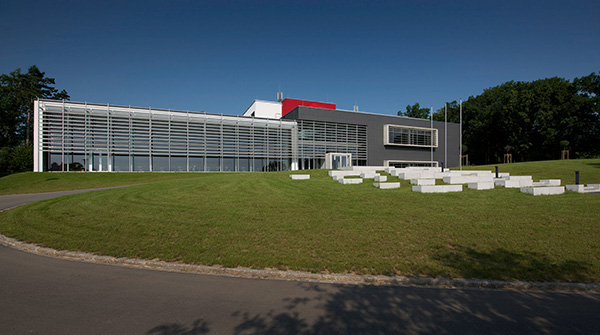 Vor dem klaren blauen Himmel der Hochschule Coburg erhebt sich ein modernes Gebäude mit Glasfassade und geometrischem Design. Es ist von einer grünen Wiese umgeben, im Vordergrund stehen weiße Steinbänke, im Hintergrund Bäume, die eine harmonische Verbindung aus Natur und Innovation schaffen.