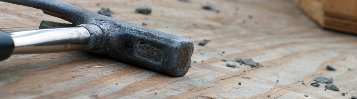 Nahaufnahme eines Metallhammers, der auf einer Holzoberfläche mit verstreuten Schuttstücken liegt. Der Hammer, der an Werkzeuge aus den Werkstätten der Hochschule Coburg erinnert, hat einen glänzenden Griff und einen verwitterten Kopf, was auf häufigen Gebrauch hindeutet. Der unscharfe Hintergrund betont die Textur des Holzes und der Schuttstücke.