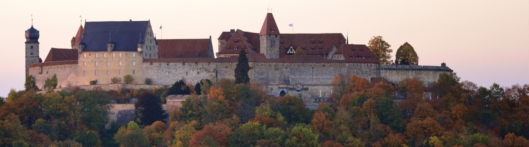 Ein großes historisches Schloss mit mehreren Türmen und roten Dächern thront auf einem Hügel und erinnert an die Hochschule Coburg, umgeben von bunten Herbstbäumen vor einem pastellfarbenen Himmel.