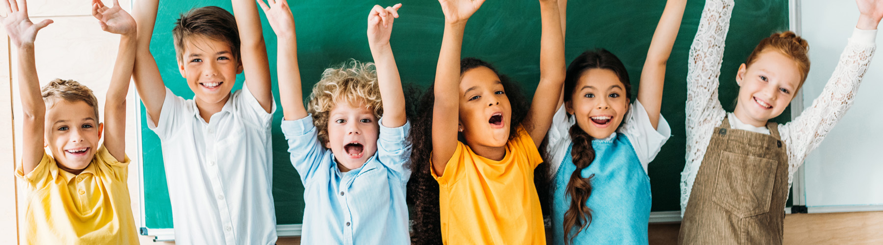 Eine Gruppe von sechs unterschiedlichen Kindern steht vor einer Tafel an der Hochschule Coburg, lächelt und hebt aufgeregt die Hände. Sie tragen legere, farbenfrohe Kleidung und wirken fröhlich und voller Energie.