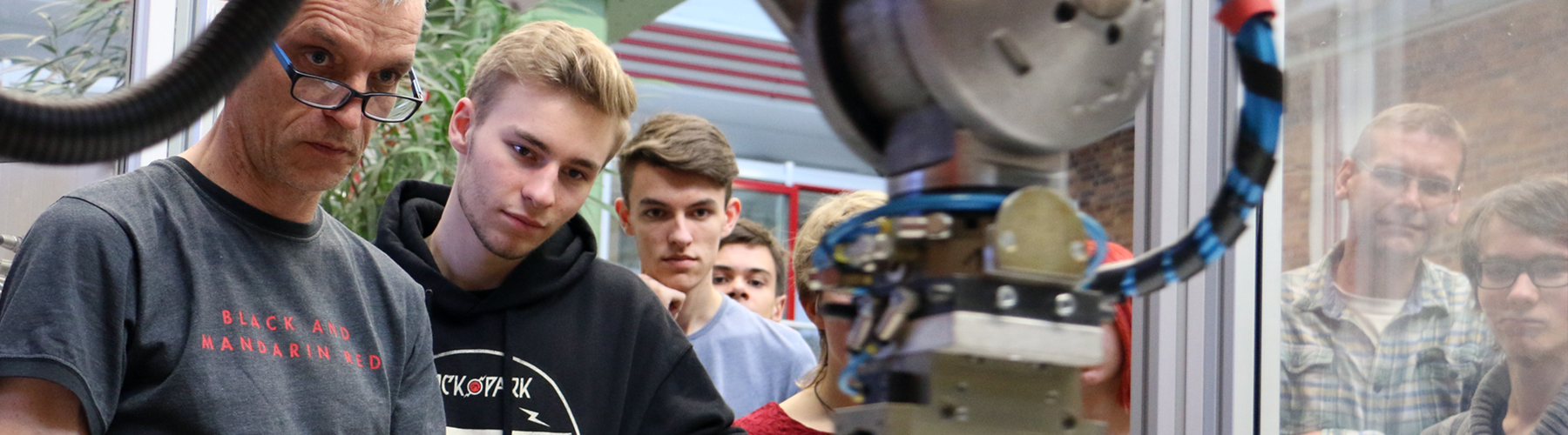 Eine Gruppe von Menschen verfolgt aufmerksam eine Vorführung eines Robotergeräts an der Hochschule Coburg. Die Szene ist eine Nahaufnahme, mit einem Mann mit Brille auf der linken Seite und mehreren jungen Erwachsenen im Hintergrund, die alle auf die Maschine blicken.