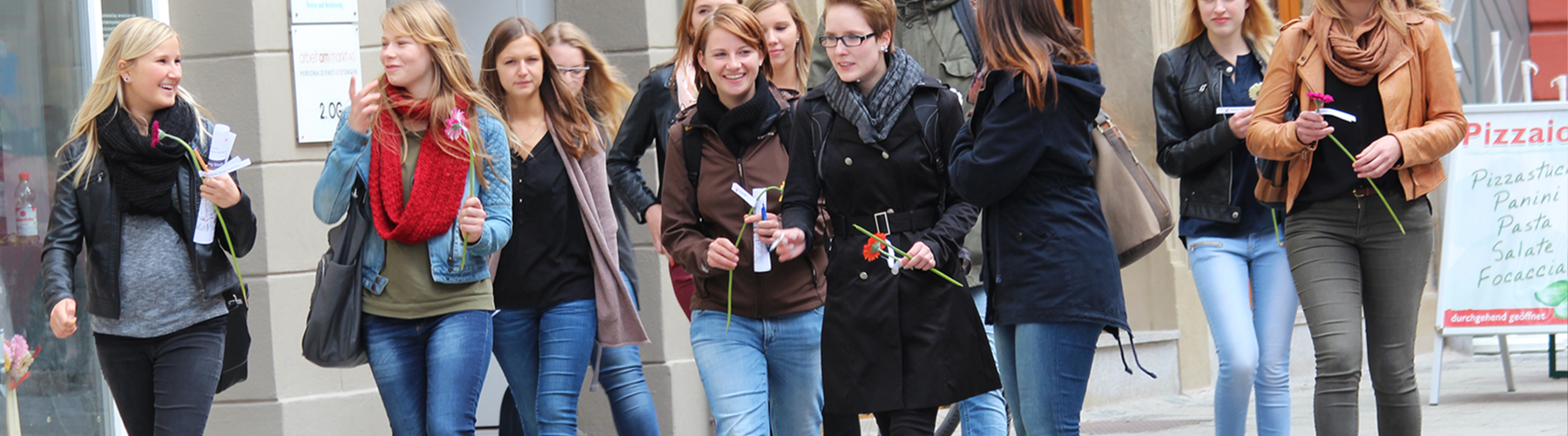 Eine Gruppe junger Frauen, vermutlich Studentinnen der Hochschule Coburg, läuft eine Stadtstraße entlang, hält Blumen in den Händen und unterhält sich. Sie sind leger gekleidet und die Atmosphäre ist lebhaft und fröhlich. Im Hintergrund ist das Schild einer Pizzeria zu sehen.