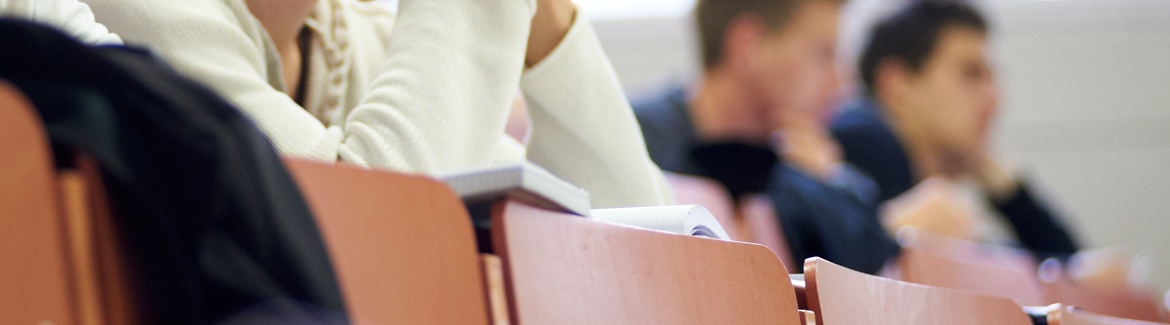 Studierende der Hochschule Coburg sitzen in einem Hörsaal mit Holzsitzen und konzentrieren sich aufmerksam auf die Präsentation. Das Bild zeigt sowohl männliche als auch weibliche Studierende mit Notizbüchern und Papieren auf ihren Tischen und betont eher die kollektive Lernumgebung als einzelne Gesichter.