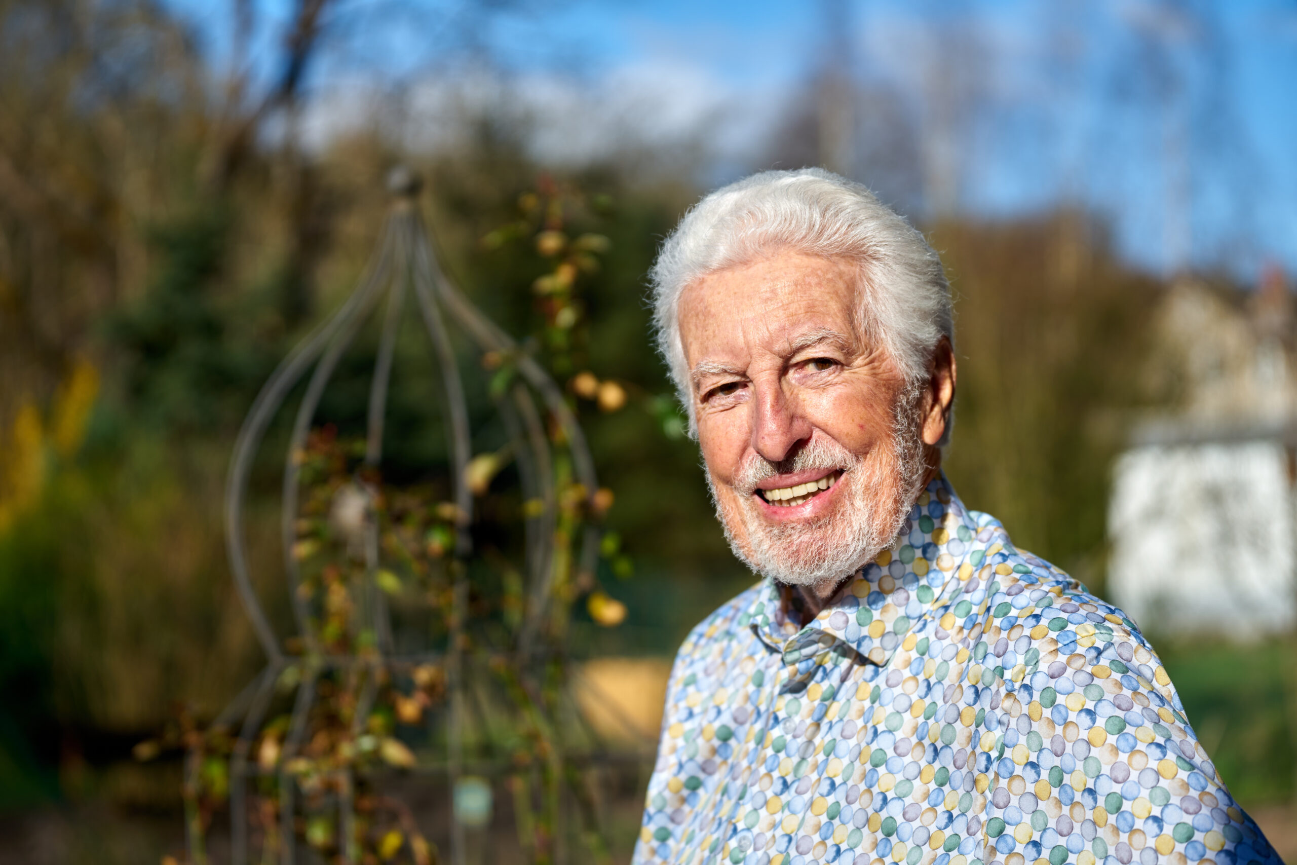 Ein älterer Mann mit weißem Haar und Bart lächelt draußen. Er trägt ein gemustertes Hemd und steht neben einer dekorativen Gartenstruktur auf dem Campus der Hochschule Coburg. Im Hintergrund sind Bäume und ein Gebäude unter einem klaren blauen Himmel zu erkennen.