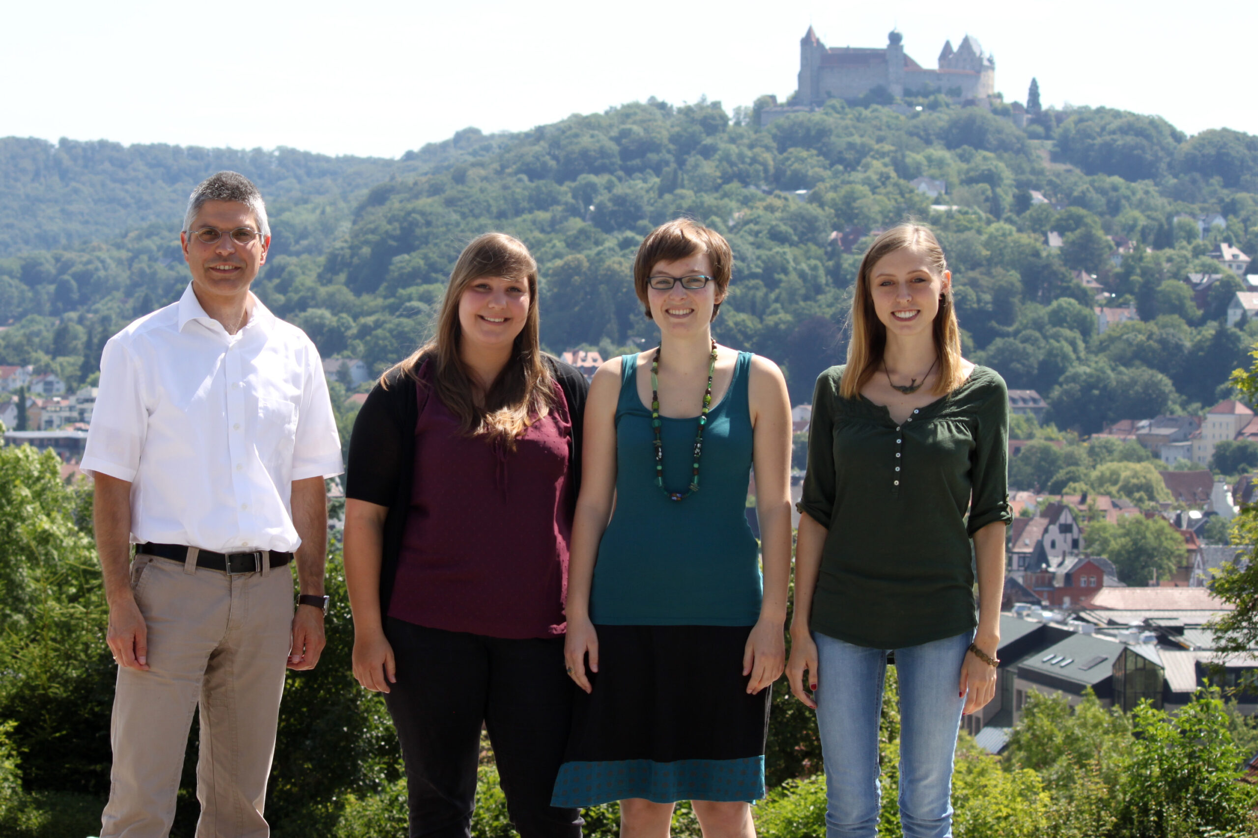 Vier Personen stehen im Freien mit einer malerischen Aussicht auf die Hügel und die historische Hochschule Coburg im Hintergrund. Sie lächeln, sind leger gekleidet und genießen einen sonnigen, klaren Tag.