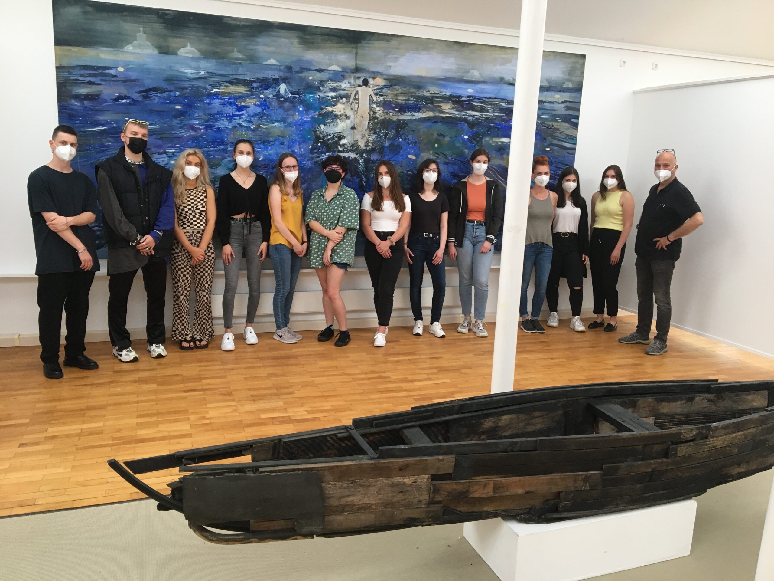 Eine Gruppe von Menschen mit Mundschutz steht in der Galerie der Hochschule Coburg. Hinter ihnen hängt ein großes Gemälde mit einer Meeresszene an der Wand. Im Vordergrund ist eine Kunstskulptur ausgestellt, die an ein Boot erinnert.