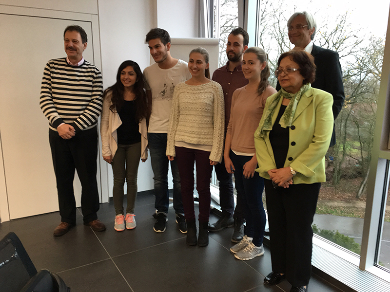 Eine Gruppe von acht Personen steht in einem Raum mit großen Fenstern der Hochschule Coburg zusammen. Einige lächeln, andere sind leger gekleidet. Zwei ältere Erwachsene stehen an den Seiten, während sich sechs jüngere Erwachsene in der Mitte versammeln. Durch die Fenster sind Bäume zu sehen.