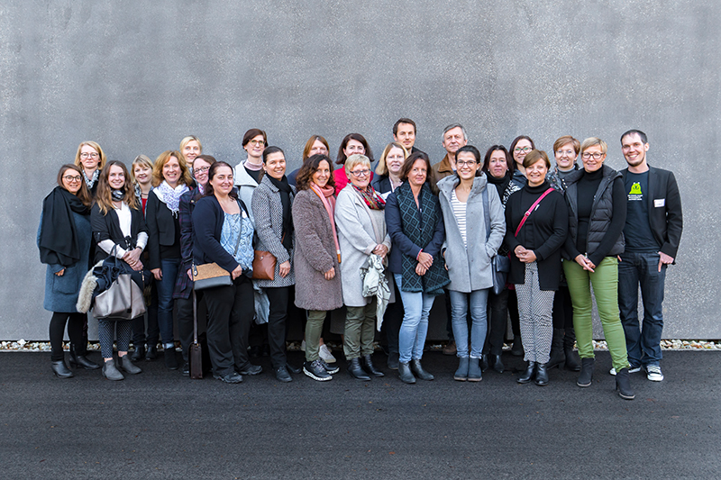 Eine Gruppe von 24 Personen, darunter sowohl Männer als auch Frauen, steht vor einer grauen Wand der Hochschule Coburg. In Freizeit- und Business-Casual-Kleidung lächeln sie in die Kamera. Der Boden ist gepflastert, einige tragen Schals und Jacken, um sich vor der Kälte zu schützen.