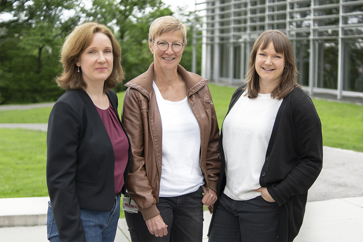 Drei Frauen stehen draußen und lächeln herzlich. Sie tragen legere, professionelle Kleidung und stehen vor der eleganten Kulisse des modernen Gebäudes der Hochschule Coburg mit seinem markanten Glas- und Metalldesign, umgeben von üppigem Grün.