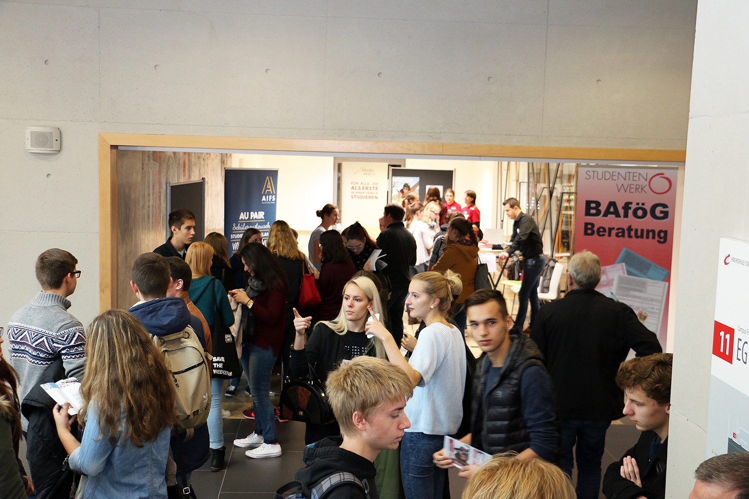 In der Halle der Bildungsmesse herrscht reges Treiben. An den Ständen werden Banner gezeigt, darunter eines für „BAföG-Beratung“, während die Besucher Informationen suchen und Broschüren mitnehmen. Unter ihnen zieht ein prominenter Stand der Hochschule Coburg neugierige Studenten an, die mehr über ihre Möglichkeiten erfahren möchten.