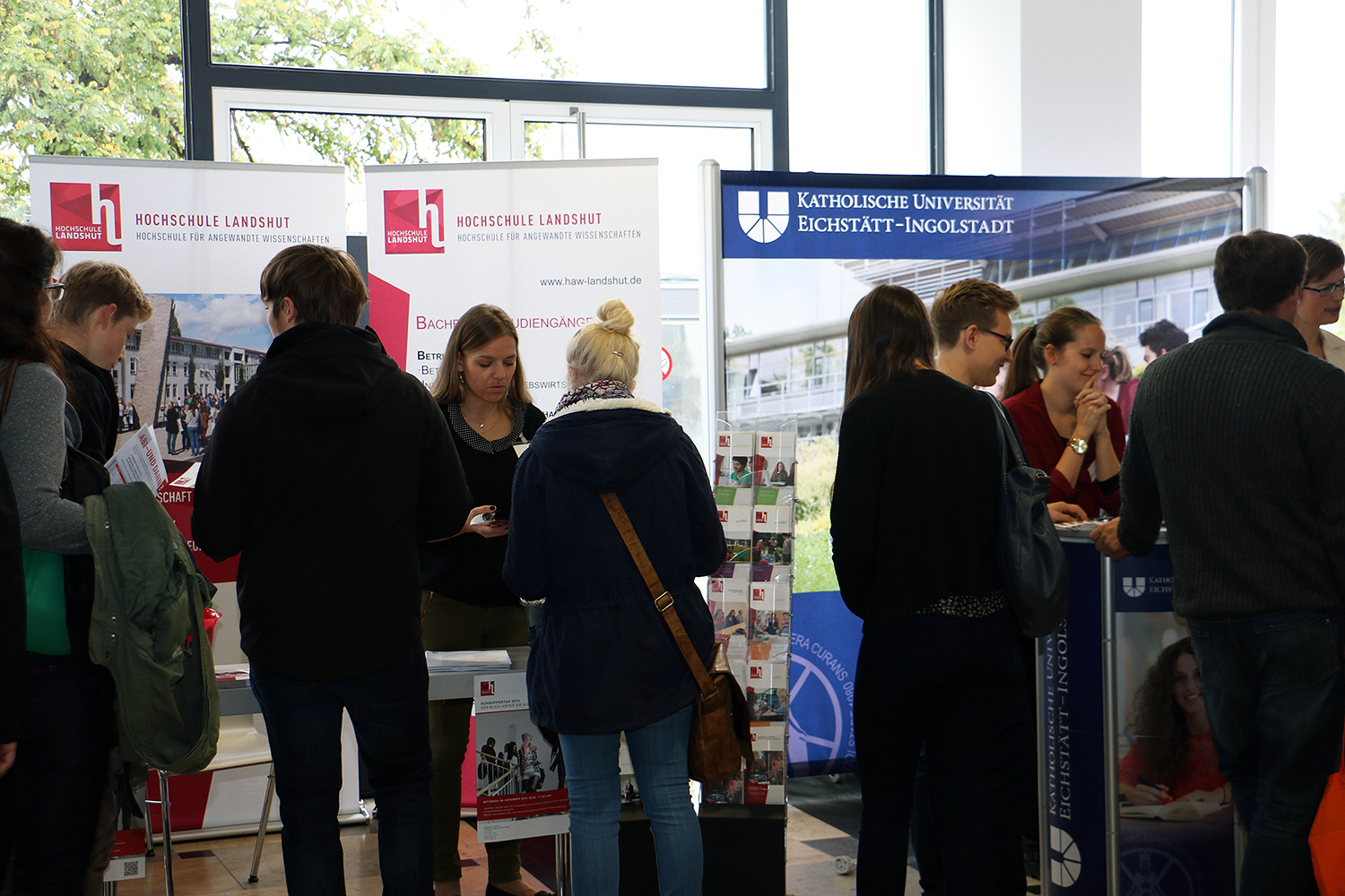 Menschen treffen sich auf einer Hochschulmesse, tauschen sich mit Vertretern aus und erkunden Informationsstände der Hochschule Landshut, der Hochschule Coburg und der Katholischen Universität Eichstätt-Ingolstadt. Auf den Tischen liegen Banner und Broschüren aus.