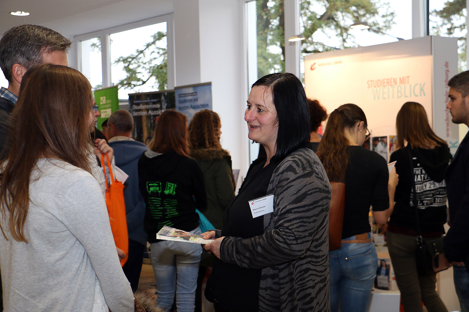 Eine Frau mit Namensschild spricht bei einer Veranstaltung in der Hochschule Coburg zu einer Gruppe. Informationsstände und Banner sorgen für eine geschäftige, gesellige Atmosphäre.