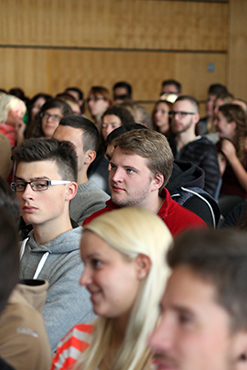 In einem Hörsaal der Hochschule Coburg sitzt eine Gruppe von Menschen eng beieinander und hört mit gemischten Gesichtsausdrücken aufmerksam zu. Im Hintergrund ist eine Holzvertäfelung zu sehen, die an eine Vorlesung oder Präsentation erinnert.