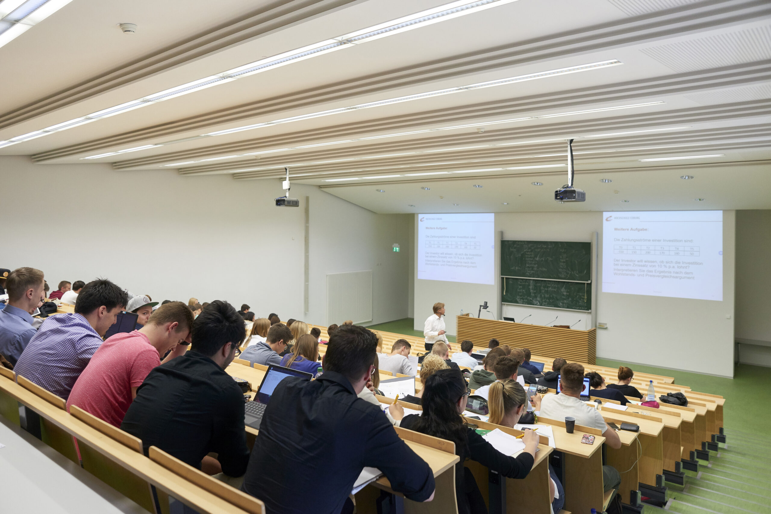 Der Hörsaal der Hochschule Coburg ist voller Studenten, die in Tischreihen sitzen und aufmerksam nach vorne blicken, wo ein Dozent einen Vortrag hält. Auf zwei Projektionsflächen und einer großen Tafel werden Notizen und Diagramme angezeigt, was das akademische Erlebnis noch bereichert.