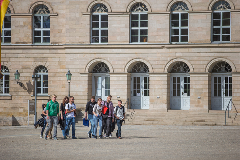 Eine Gruppe junger Leute schlendert gemeinsam vor dem historischen Gebäude der Hochschule Coburg, das durch seine Rundbogenfenster und zahlreichen Türen charakterisiert ist. Lässig gekleidet unterhalten sie sich angeregt unter dem sonnigen Himmel.