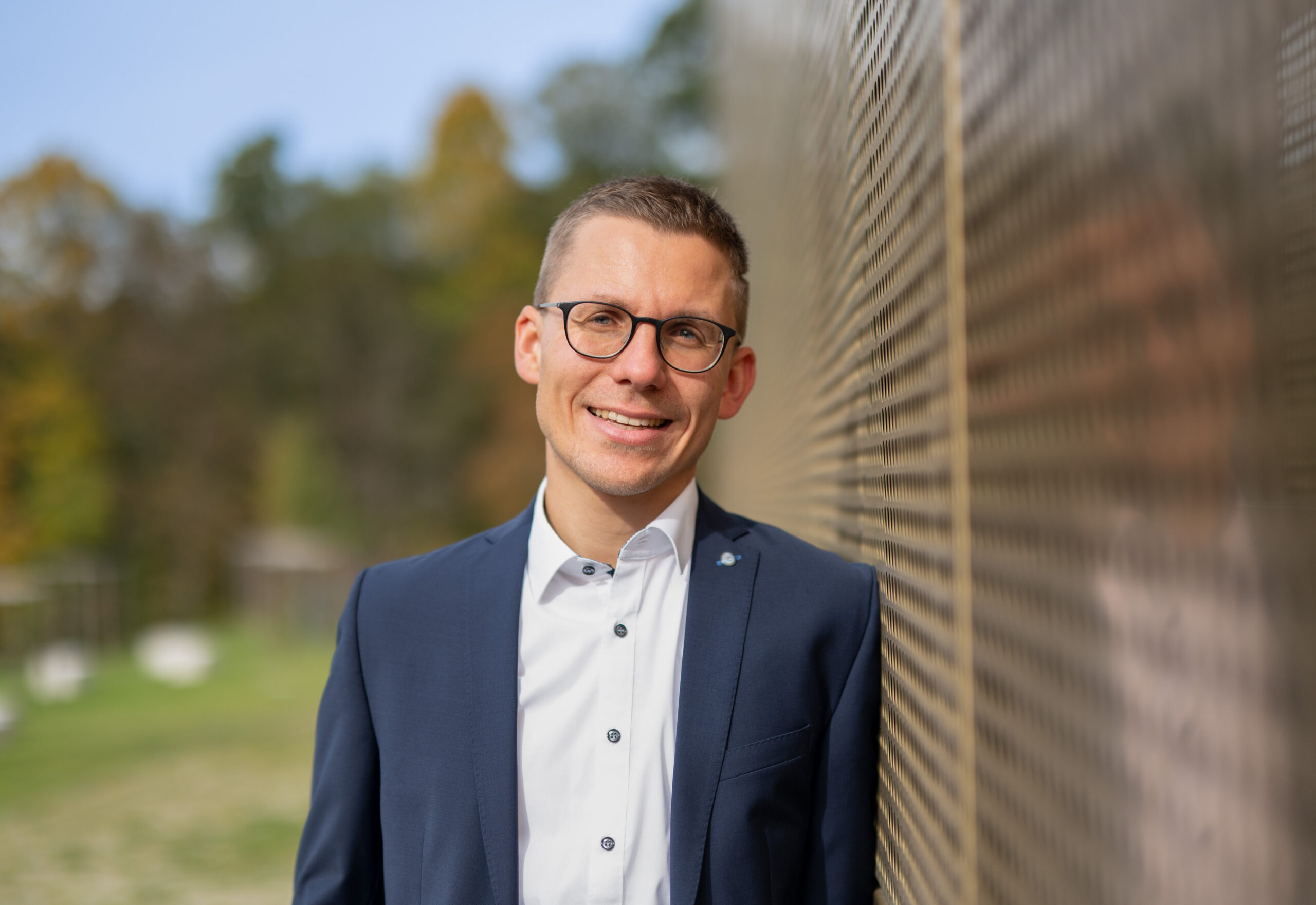 Ein Mann in blauem Anzug und weißem Hemd lehnt draußen an einer reflektierenden Wand der Hochschule Coburg. Er lächelt unter seiner Brille und steht vor einem verschwommenen Hintergrund aus Bäumen und Grün unter einem klaren Himmel.