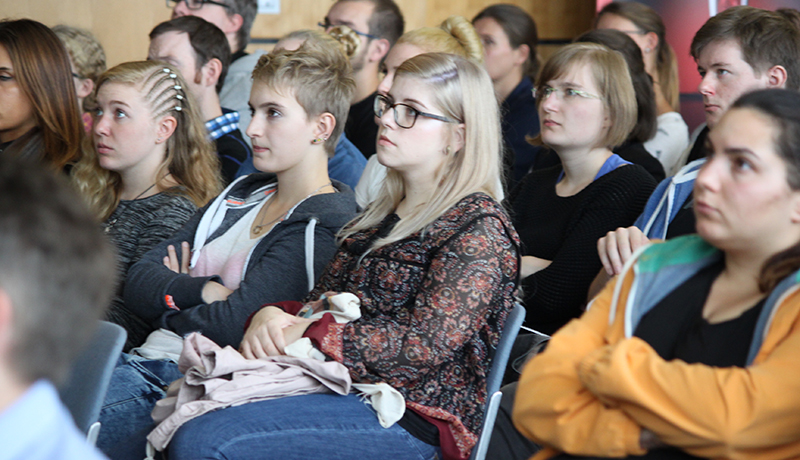 Eine bunt gemischte Gruppe von Menschen sitzt aufmerksam in einem Hörsaal der Hochschule Coburg und hört sich einen Vortrag an. Die meisten tragen legere Kleidung und Brillen, ihre Miene ist konzentriert. Der Raum strahlt eine lockere, akademische Atmosphäre aus.