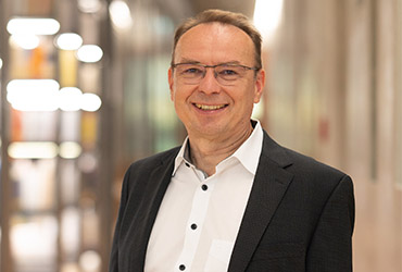 A smiling man with short hair and glasses stands in a corridor adorned with blurred lights, wearing a dark suit jacket and white shirt, embodying the professionalism of Hochschule Coburg.