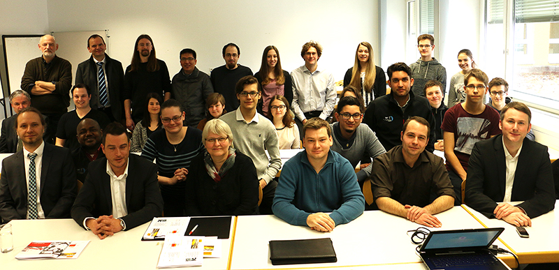 Eine Gruppe von Menschen posiert in einem Unterrichtsraum der Hochschule Coburg. Einige sitzen an Tischen, andere stehen hinter ihnen. Der gut beleuchtete Raum strahlt eine lockere, professionelle Atmosphäre aus. Auf den Tischen liegen verstreut Laptops und Dokumente.