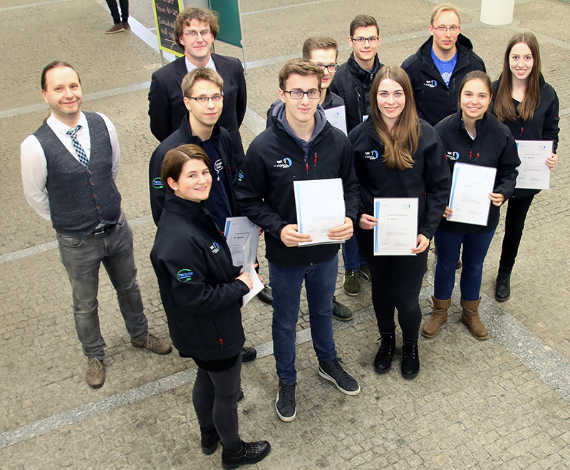 Eine Gruppe von Menschen steht im Innenbereich der Hochschule Coburg, einige halten Zertifikate in den Händen. Sie tragen Jacken und lächeln in die Kamera, um die Essenz dieses offiziellen akademischen Ereignisses einzufangen.