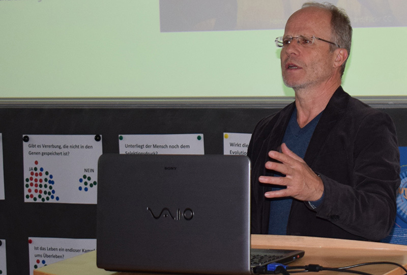 Eine Person mit Brille hält hinter einem Laptop auf einem Podium der Hochschule Coburg eine Präsentation. Im Hintergrund sind Plakate mit verschiedenen Texten auf Deutsch zu sehen, während auf dem aufgeklappten Laptop stolz der Markenname prangt.