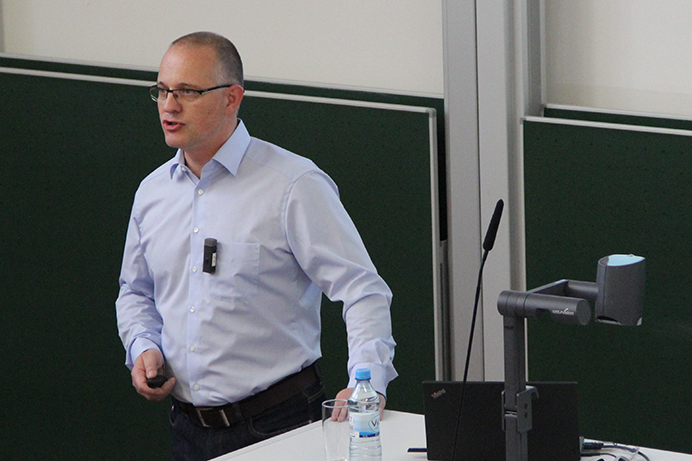 Eine Person mit Brille und hellblauem Hemd hält in einem Hörsaal der Hochschule Coburg einen Vortrag. Sie steht mit einem Laptop und einer Flasche Wasser neben einem Podium, während hinter ihr teilweise eine Projektionsfläche zu sehen ist.