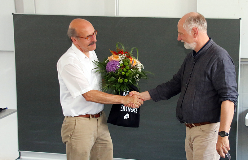 Zwei Männer schütteln sich vor einer Tafel die Hände, einer überreicht dem anderen eine Geschenktüte mit Blumen. Sie sind leger gekleidet und scheinen sich in einem Unterrichtsraum oder einer Vorlesung an der Hochschule Coburg zu befinden.