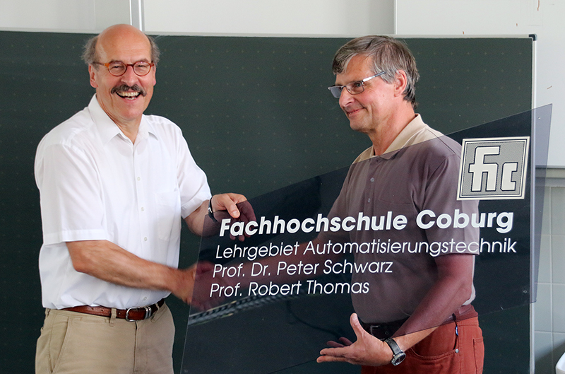 Zwei Männer lächeln und halten ein transparentes Schild mit dem Text „Hochschule Coburg“, dem Namen „Fachhochschule Coburg“ und Angaben zum „Lehrgebiet Automatisierungstechnik“ mit den Namen zweier Professoren hoch. Sie stehen vor einer Tafel.