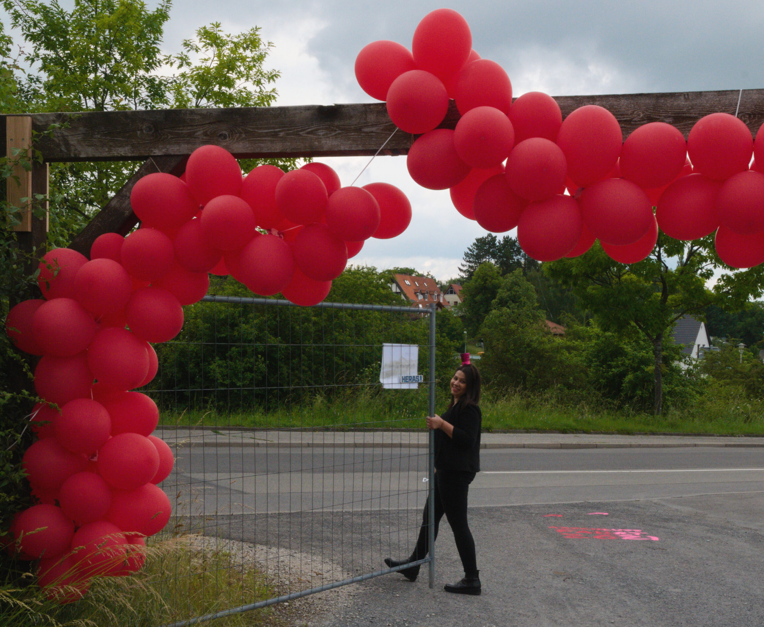 Eine Person steht lächelnd neben einem Torbogen aus roten Luftballons an einem leicht bewölkten Tag und feiert in der Nähe der Hochschule Coburg. Die Luftballons sind elegant an einer Holzkonstruktion am Straßenrand befestigt, im Hintergrund sind Bäume und Häuser zu sehen.